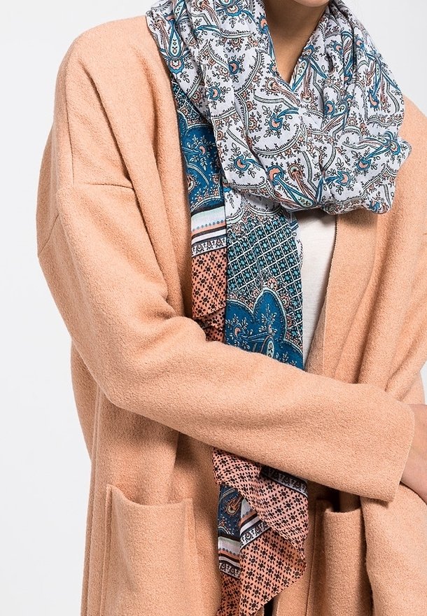 Personne portant un manteau en laine beige et une écharpe à motifs avec des dessins paisley et floraux bleus, blancs et orange.