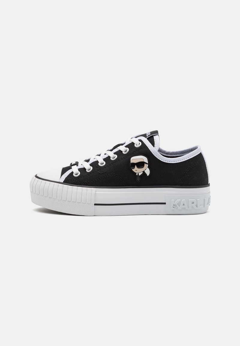 Black canvas sneakers with white accents, round toe, textured platform sole, featuring a cartoon character pin on the side and logo detail.