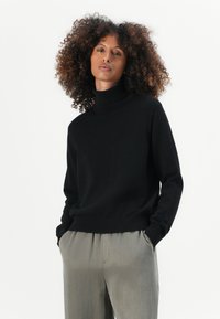 Pull sous-pull noir en laine douce avec des poignets et un ourlet côtelés, associé à un pantalon gris clair, sur fond blanc uni.