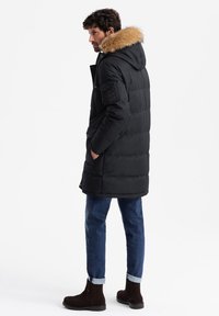 Cappotto imbacuccato nero con cappuccio foderato in pelliccia, dotato di zip frontale, tasche laterali e una toppa testurizzata sulla manica. Indossato con jeans blu e stivali marroni.