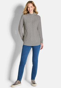 Grauer Strickpullover mit einem Zopfmuster, hohem Kragen und geripptem Saum, kombiniert mit blauen Jeans und metallischen Schuhen.
