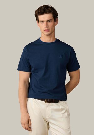 Joven con cabello rizado y castaño que lleva una camiseta de manga corta azul marino y pantalones beige con un cinturón tejido marrón sobre un fondo liso.