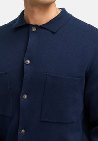 Navyblaues, strukturiertes Strickjacke mit Kragen, zwei Brusttaschen und fünf Knöpfen entlang der Vorderseite.