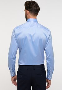Chemise bleu clair avec une texture lisse, des manches longues, des poignets à boutons et un design de découpe au dos. Assortie à un pantalon sombre.