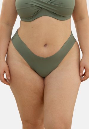 Plus-Size-Frau trägt olivgrüne Bikinihose und Oberteil, gezeigt vom mittleren Oberschenkel bis unter die Brust, mit entspannten Armen an den Seiten.