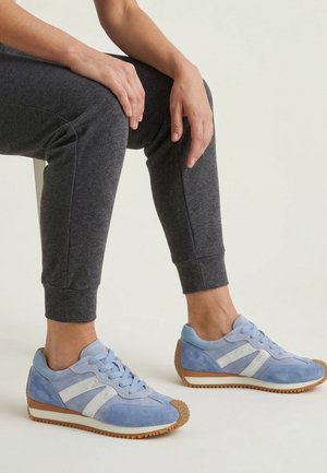 Person sitzt, trägt graue Jogginghosen und hellblaue Sneakers mit weißen Akzenten und Gummisohlen vor einem schlichten hellen Hintergrund.