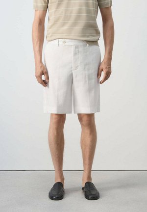 Mann trägt beige gestreiftes Poloshirt, knielange weiße Shorts und schwarze Slipper, steht vor einem schlichten hellen Hintergrund.
