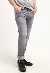 Vaqueros de mezclilla gris con corte ajustado, bajos enrollados y bolsillos frontales. Combinados con zapatillas de lona negras con cierre de cordones y suelas de goma.