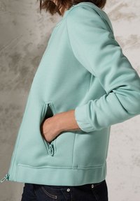 Osoba v svetle tyrkysové zipové bundě, jejíž ruka je částečně zasunuta v zipové postranní kapse, na texturovaném šedém pozadí.