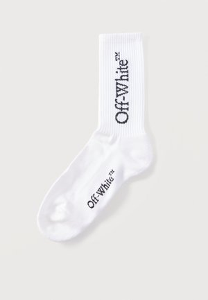 Balta kojinė su juosta ir juodu „Off-White™“ logotipu, spausdintu vertikaliai ant juostos ir horizontaliai šalia kojų pirštų.