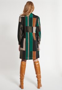 Robe à manches longues présentant un motif géométrique en vert, marron et noir. Tissu lisse avec une coupe décontractée et des détails de boutons aux manches.