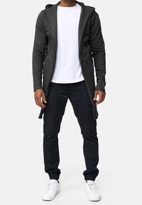 Sudadera larga de color gris oscuro con cremallera, con un corte relajado, capucha grande y detalles de aberturas laterales, usada sobre una camisa blanca y pantalones negros casuales.