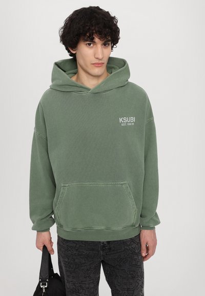 Zielona bluza z kapturem, wyposażona w przednią kieszeń i haftowane logo, zestawiona z wzorzystymi czarnymi dżinsami oraz czarną torbą. Luźny, swobodny krój.