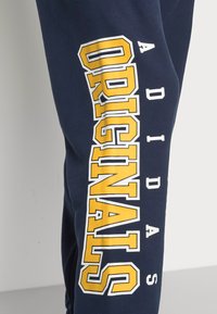 Tumši zila kokvilnas sporta bikses ar izteiksmīgu dzeltenu un baltu tekstu "ADIDAS ORIGINALS" sānos sportiskā fontā.
