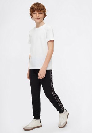 Junge mit kurzen lockigen Haaren, der ein weißes T-Shirt, schwarze Jogginghosen mit Logostreifen und weiße Sneaker trägt, steht vor einem schlichten weißen Hintergrund.