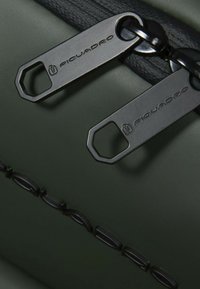 Bolsa verde oscuro con superficie texturizada, tiradores de cremallera negros con la marca "Piquadro" y detalles circulares a lo largo del lateral.
