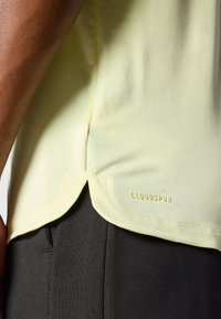 Lys gul sports skjorte med rundet kant, sidespalter og et "CLOUDSPUN" mærke. Kombineret med mørkegrå bukser.
