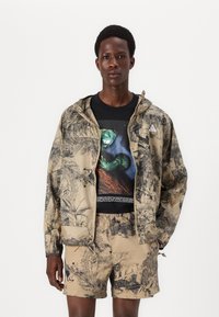 Leichte Kapuzenjacke in Beige mit schwarzen, von der Natur inspirierten Drucken, kombiniert mit einem schwarzen Grafik-T-Shirt und passenden Shorts.