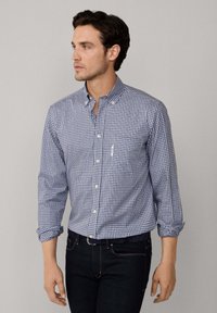 Hombre vestido con una camisa de cuadros azul y blanca de botones, con las mangas arremangadas y jeans oscuros, mirando hacia su izquierda frente a un fondo sencillo.