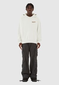 Weißer Kapuzenpullover mit einer vorderen Tasche und Logo, kombiniert mit schwarzen Cargo-Hosen, die mehrere Taschen und einen lockeren Schnitt haben.
