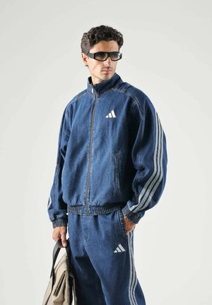 Uomo con tuta Adidas in denim blu scuro e occhiali neri che tiene una borsa beige su uno sfondo chiaro semplice.
