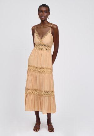 Maxi-jurk van lichtbeige stof met een diepe V-hals, kanten details op het lijfje en de lagen, en dunne schouderbandjes. Bruine sandalen.