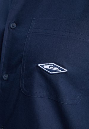 Granatowa koszula zapinana na guziki z kieszonką z przodu i haftowanym białym logo, prezentująca elegancką fakturę oraz dopasowany fason.