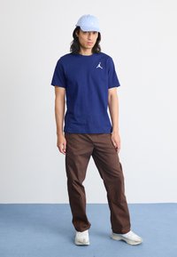 Bărbat îmbrăcat cu o pălărie albastră, un tricou Jordan bleumarin, pantaloni maro și adidași albi, stând pe un podea albastră, în fața unui perete alb.