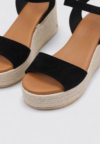 Sandalias de cuña de ante negro con suela de yute, que presentan una tira ancha sobre los dedos del pie y una correa ajustable en el tobillo. Plantilla de cuero suave.