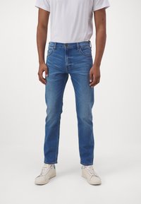Blaue Jeans aus Denim mit geradem Schnitt, fünf Taschen und klassischem Knopfverschluss. Getragen mit einem schlichten weißen T-Shirt und hellen Sneakers.
