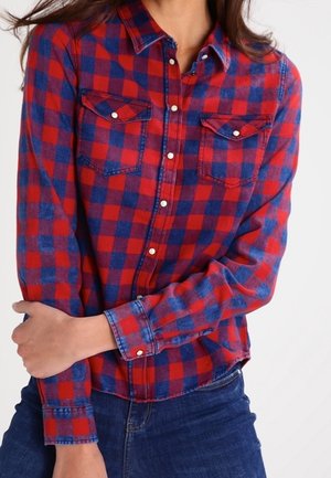 Femme portant une chemise à carreaux rouge et bleu à manches longues et un jean bleu, bras croisés à la taille.