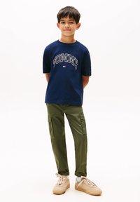 Tricou din bumbac de culoare albastru marin cu logo-ul "TOMMY", pantaloni cargo de culoare verde măsliniu cu buzunare laterale, adidași bej și șosete albe.