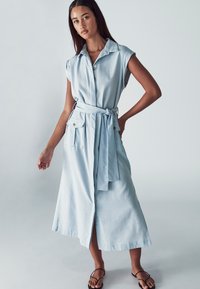 Vaik-blue denim-mekko, jossa on lyhyet hihat, kaulus, etunappit, kaksi sivutaskua ja vyötärölle sidottava vyö. Tasainen rakenne ja midi-pituus.
