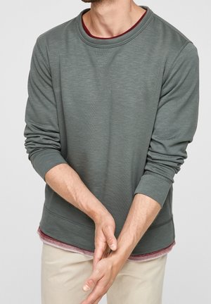 Mand iført grøn langærmet skjorte over en rød undertrøje og beige bukser, med hænderne foldet foran sig mod en neutral baggrund.