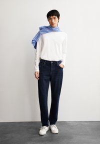 Witte blouse met lange mouwen, donkerblauwe spijkerbroek en witte schoenen. Een blauw-wit gestreepte sjaal hangt over de schouder. Soepele textuur.