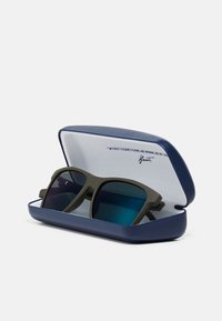 Óculos de sol matte verde oliva com lentes azul-tintadas dentro de um estojo rígido azul-marinho aberto com revestimento interior branco.