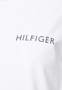 Bílý bavlněný tričko s krátkými rukávy, které má na sobě nápis "HILFIGER" z černých kamínků, umístěný mírně mimo střed. Hladký povrch.