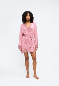 Γυναίκα που στέκεται ξυπόλητη φορώντας ροζ σατέν ρόμπα με δαντελωτή περίμετρο και ταιριαστό δαντελωτό κορμάκι από κάτω σε απλό φόντο.