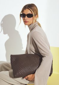 Brun vävd läderclutch med en böjd form. Modell bär en ljusbeige polotröja och håller väskan mot en ljus bakgrund.