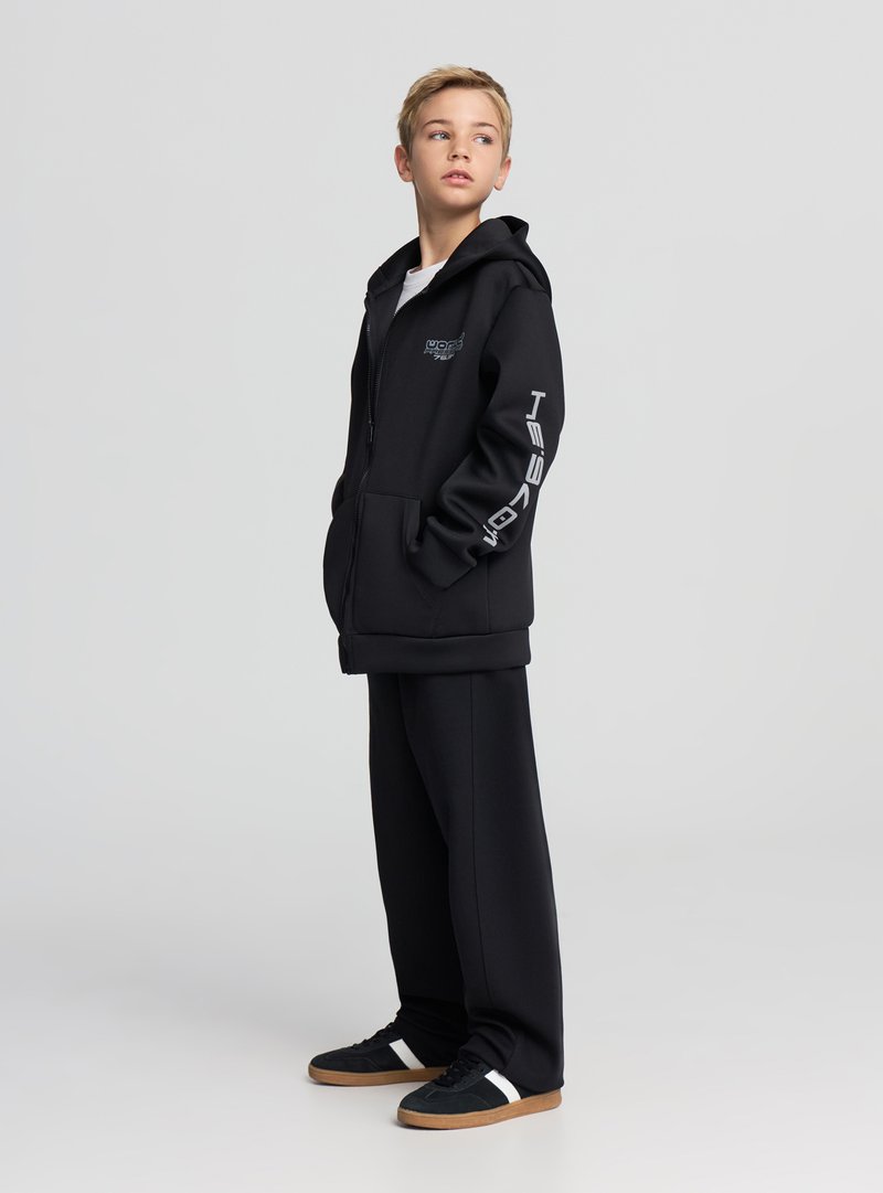 Felpa nera con zip, due tasche frontali, logo argentato sul petto e design con testo sulla manica. Abbinata a pantaloni neri e sneakers.