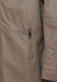 Braune Lederjacke mit einer seitlichen Reißverschlusstasche, glatter Textur und dezenten Nähten am Saum. Logo an der Unterseite eingestickt.