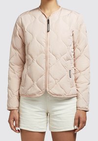 Woman wearing a light pink quilted zip-up jacket with a rounded neckline, paired with light beige shorts against a plain background.