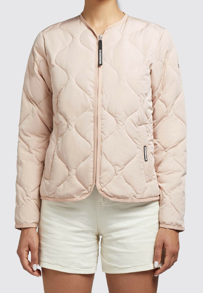 Woman wearing a light pink quilted zip-up jacket with a rounded neckline, paired with light beige shorts against a plain background.