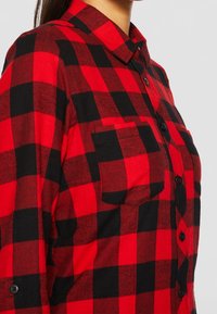 Femme portant une chemise en flanelle à carreaux rouges et noirs avec des boutons et un col noirs, vue de l’épaule à la poitrine sur un fond uni.
