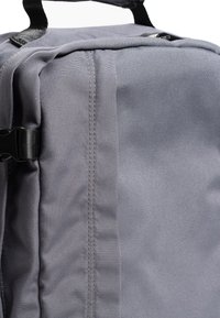 Šedý látkový batoh s odolnou textilií, detailním prošitím a boční sponou pro bezpečné uzavření. Minimalistický design s hladkou texturou.