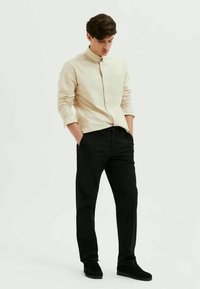 Camisa bege de botão com mangas longas, combinada com calças pretas e sapatos escuros. A camisa tem colarinho e um design ajustado.