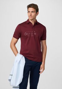 Burgundské polo tričko s knoflíkovým límcem a jemným logem. Spárované s námořnickými kalhotami a světlomodrou bundou přehodenou přes jedno rameno.