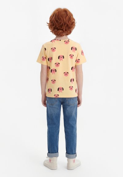 Kind met krullend rood haar, gekleed in een geel T-shirt met roze hondenkoppen, blauwe opgerolde jeans en witte hoge sneakers met roze sterren.