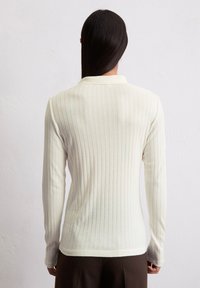 Langärmliges, eng anliegendes Oberteil in Off-White mit vertikalen Rippungen. Verfügt über einen klassischen Kragen und eine weiche Textur, kombiniert mit dunkelbraunen Hosen.
