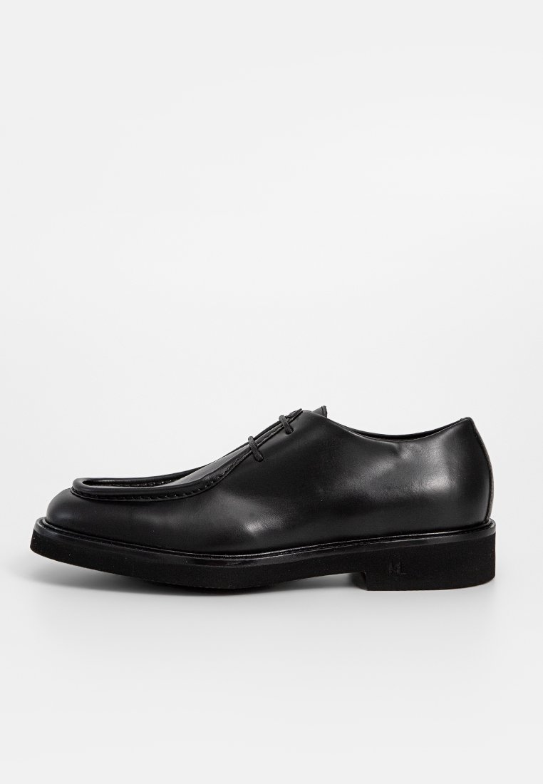 Zapato de vestir de cuero negro con punta redondeada, costuras decorativas y suela de goma plana. Presenta un diseño único doblado en el empeine.